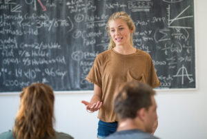 Female student talking at front of class with math equations on chalkboard.