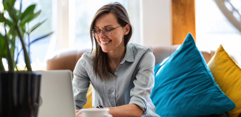 Woman sitting on sofa with blue and yellow pillows smiling at laptop.