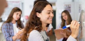 Female student smiling while writing with blue marker on white board.