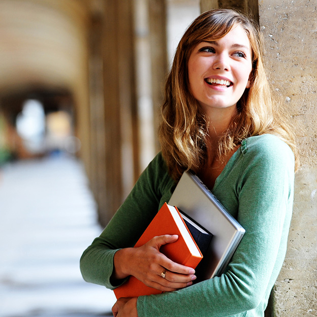 Smiling female college student leaning against stone wall holding books and contemplating her education and career pathway