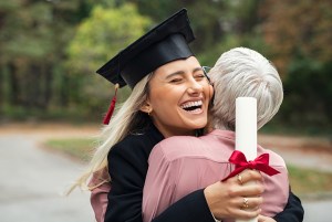 Female Graduate Hugging older woman while wearing cap and gown holding diploma.