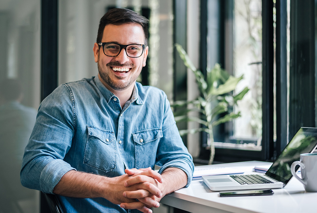 Man in denim shirt sitting at desk with laptop smiling.