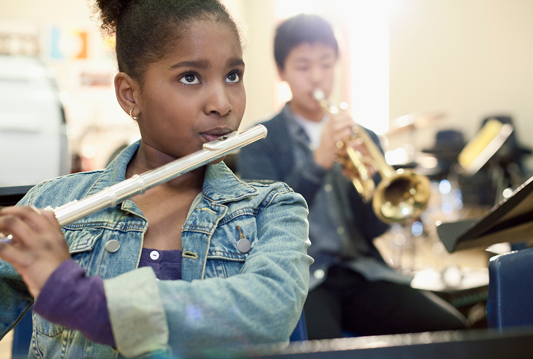 Young female student practicing the flute and young male student practicing the trumpet sitting in the background.