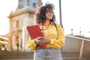 Woman with curly hair wearing yellow shirt and glasses holding notebooks.