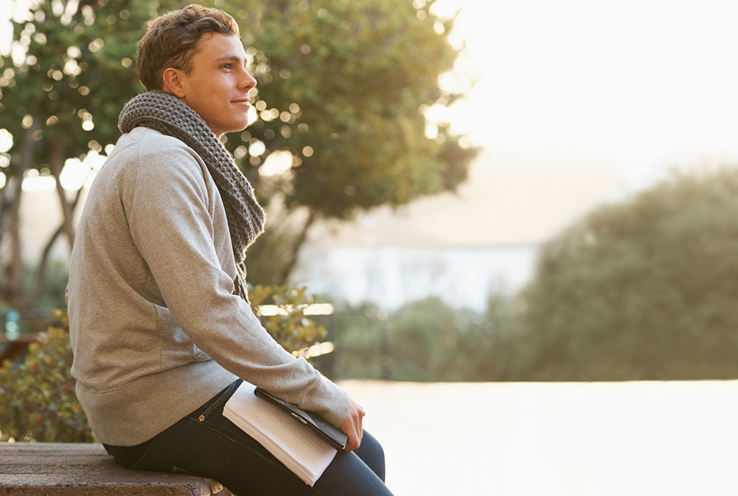 Male student wearing scar, holding books sitting on stone wall.