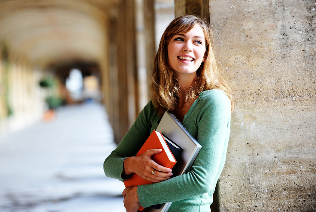Female in green sweater holding books leaning against cement pillar.
