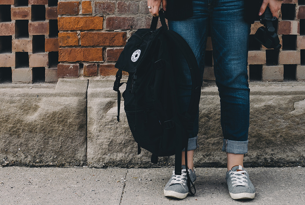 Black backpack held by a woman standing in front of a brick wall.