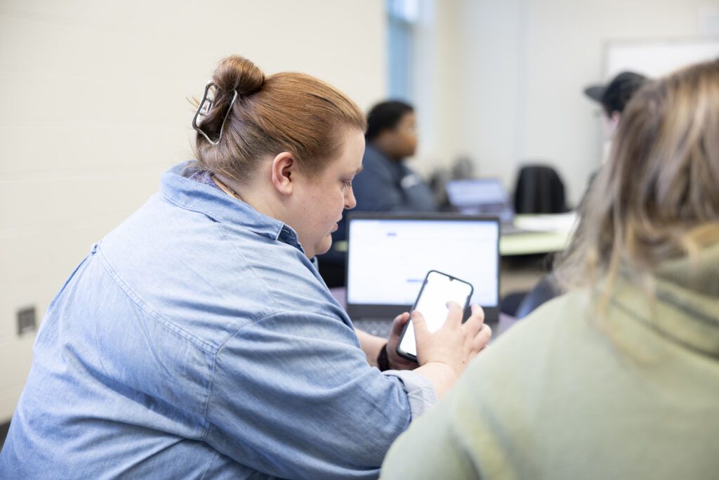 Two women sitting at a classroom table, viewing something on one woman's phone.