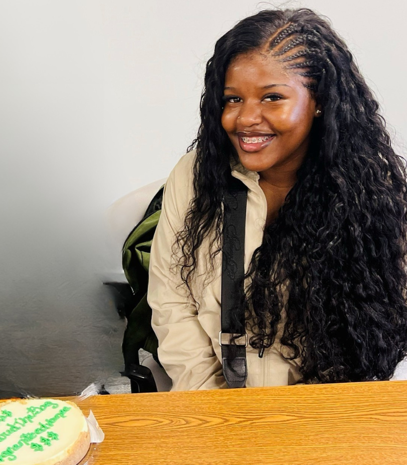 A young woman sits at a table that holds a cheesecake.